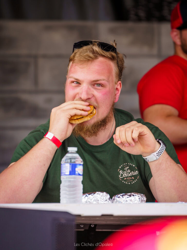 Homme dégustant un burger lors d'un événement en extérieur, portant un t-shirt vert et une montre, a.