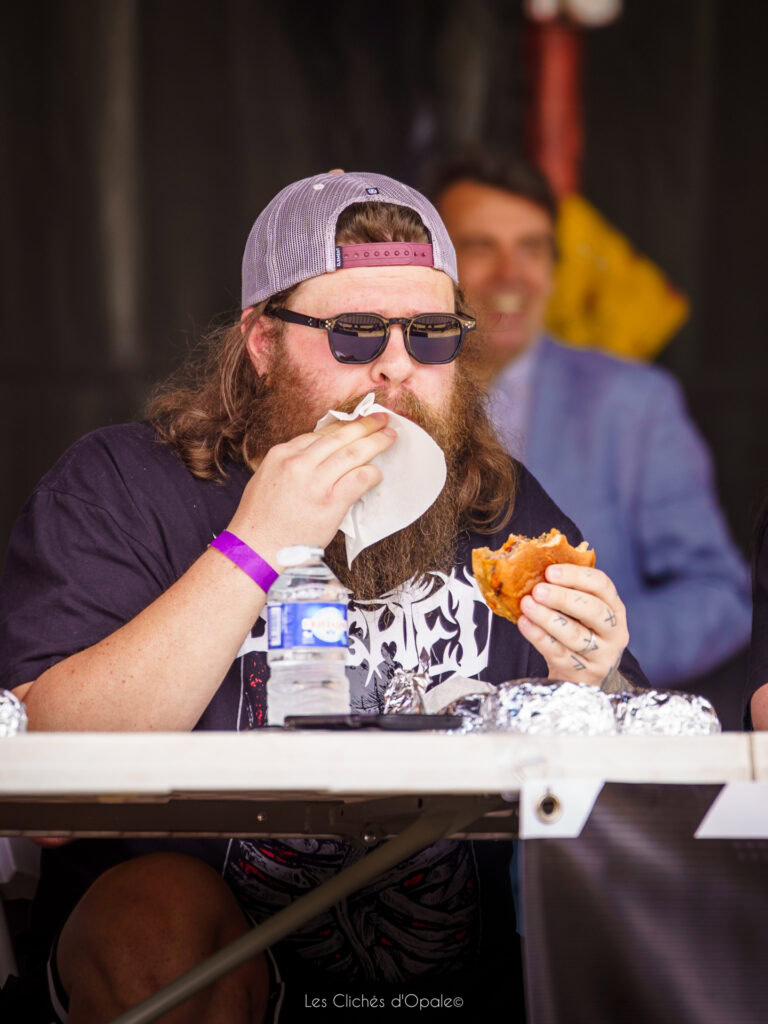 Homme avec barbe, lunettes de soleil, chapeau, dégustant une pizza dans un cadre décontracté.